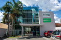 Fachada de um prédio moderno com grande painel de vidro espelhado. À direita, na parede branca, destacam-se placas do "Cadastro Único" e da "DPE-PR em Cianorte". Há palmeiras e carros estacionados na entrada.