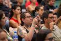 Imagem mostra detalhe do público presente. Em primeiro plano uma mulher com tatuagens registra o evento com celular. 
