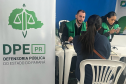 Close-up de um atendimento onde um servidor de colete verde conversa com uma mulher sentada de costas. Ao lado dele, outra profissional manuseia o celular, com um banner da DPE-PR em grande destaque no lado esquerdo.