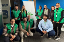 Dez profissionais da equipe da DPE-PR posam sorridentes para uma foto em grupo. A maioria veste coletes verdes da instituição, dividindo-se entre pessoas em pé e agachadas ao redor de um banner institucional.