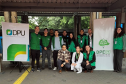 Uma equipe de doze pessoas posa para foto em um espaço público com piso tátil amarelo no chão. A equipe, com vários membros usando coletes verdes, está posicionada entre os banners da Defensoria Pública da União (DPU) e da DPE-PR.