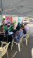 Em uma tenda de atendimento na rua a equipe da Defensoria Pública conversa e orienta as pessoas sentadas à frente das mesas. Ao fundo, destaca-se um banner branco e verde da DPE-PR.