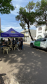 Uma tenda azul com a inscrição "Centro da Juventude" montada na rua abriga pessoas em atendimento. Estacionada logo ao lado, sob a sombra de uma árvore, está uma caminhonete branca com a logo verde da DPE-PR.