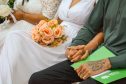 Close no colo de um casal sentado. A noiva, de vestido branco, segura um buquê de rosas pêssego, enquanto o companheiro ao lado apoia as mãos, uma delas tatuada e com aliança, sobre uma pasta verde da DPE-PR.