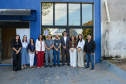 Um grupo de dez pessoas posa sorridente para uma fotografia em frente à fachada azul e às portas de vidro de um prédio identificado como "COMPLEXO SOCIAL". A equipe veste trajes sociais casuais, e alguns usam crachás de identificação no pescoço.