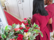 Um casal, com a mulher vestindo um vestido vermelho, caminha por um corredor coberto por um tapete vermelho. Em primeiro plano e ligeiramente desfocados, arranjos de flores brancas, rosas e vermelhas enfeitam a lateral do caminho.