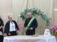 Um celebrante com uma faixa verde e amarela no peito está posicionado atrás de uma mesa com um pequeno bolo de casamento enfeitado com flores. Ao lado dele, uma mulher de blusa preta e branca acompanha a cerimônia em frente a um arco floral.