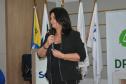 Uma mulher de cabelos escuros e conjunto preto segura um microfone enquanto fala. Logo atrás dela, estão posicionadas mastros com bandeiras oficiais e parte de um banner da Defensoria Pública.