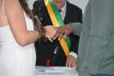 Close de um casal trocando alianças em uma cerimônia. A noiva veste branco e o noivo veste uma camisa verde. Ao fundo, o celebrante que segura o microfone usa uma faixa verde e amarela, e uma caixinha vermelha de anel repousa sobre a mesa.