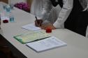 Foco nas mãos de uma pessoa de camisa branca e colete escuro assinando documentos sobre uma mesa clara. Próximo às mãos, há certidões de casamento com brasão oficial, caixinhas vermelhas para alianças e copos de água.