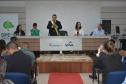 Visão ampla de um auditório onde um celebrante com faixa verde e amarela fala ao microfone em uma mesa diretiva com as logos do Fecomércio PR e Senac. À esquerda, há um banner da DPE-PR. O público assiste sentado, com pessoas fotografando a cena.