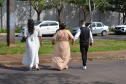 Visão de costas de três pessoas caminhando de mãos dadas por uma rua asfaltada e arborizada com carros estacionados. Da esquerda para a direita, caminham uma noiva de vestido branco, uma mulher de vestido longo bege e um noivo de colete preto e camisa branca.