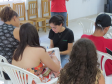 Uma jovem de camiseta preta com a logo da DPE-PR anota informações em uma prancheta enquanto atende pessoas que estão sentadas ao seu redor em cadeiras de plástico branco.