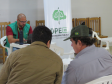 Um servidor de óculos e colete verde atende em um notebook numa mesa branca. À sua frente, dois homens aguardam sentados de costas. Ao fundo, há um banner vertical da DPE-PR.