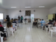 Visão ampla de um salão com diversas pessoas sentadas em cadeiras brancas voltadas para o fundo. Lá, servidores da DPE-PR estão próximos a mesas posicionadas ao lado de um banner da instituição.