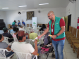 Um funcionário em pé, de óculos e colete verde, entrega um panfleto a um homem sentado de costas. Ao lado do servidor há um carrinho com um bebê dormindo, e outras pessoas aguardam ao fundo do salão.