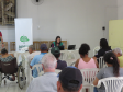 Uma mulher de colete verde trabalha em um notebook ao fundo da sala, ao lado de um banner da DPE-PR. À frente dela, diversas pessoas aguardam sentadas de costas, incluindo uma pessoa em cadeira de rodas.