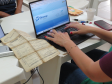 Fotografia em close de mãos femininas com esmalte vermelho digitando em um notebook que exibe a página do sistema Sinesp. Sobre a mesa branca, à esquerda do teclado, destaca-se um documento oficial muito antigo, amarelado e com marcas de dobras. Canetas, um celular e um copo descartável também compõem a cena.