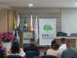 Um auditório com o público sentado de costas em cadeiras azuis, voltado para um palco. No palco, há um púlpito com a logo do Senac acompanhado por bandeiras oficiais, e à direita destaca-se um banner da Defensoria Pública (DPE-PR).