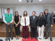 Seis pessoas posam lado a lado em um palco de piso de madeira com um tapete vermelho no centro. O grupo é formado por homens e mulheres em trajes sociais, incluindo, à extrema esquerda, um homem vestindo o colete verde da DPE-PR.