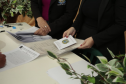 Detalhe de uma mesa com vários documentos impressos. Duas mulheres de terno preto manuseiam os papéis, e uma delas segura um livreto com o brasão da República do Brasil na capa. À esquerda, vê-se a mão de uma pessoa assinando um papel.
