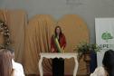 Uma mulher com blazer vermelho e a faixa transversal verde e amarela está em pé atrás da mesa branca, falando aos presentes. O cenário mantém os painéis dourados ao fundo e o banner da DPE-PR à direita.