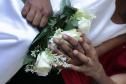 Detalhe em close de mãos negras entrelaçadas sobre um buquê de rosas brancas, ambas exibindo alianças douradas. Uma das pessoas veste um traje branco (vestido), e a outra usa uma blusa vermelha.