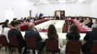 Vemos todos os participantes do encontro. A foto foi tirada atrás da mesa principal dos palestrantes, então vemos esses de costas e os participantes sentados em largas mesas laterais e uma frontal. 