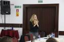 A Assistente Social do MPPR, Clarice Metzner, está de pé atrás da mesa principal para os palestrantes falando. 
