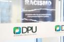 A foto foi tirada de uma porta transparente. onde está colada a logo da DPU. Essa se encontra em destaque na imagem. 