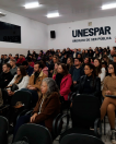 Imagem da plateia sentada prestando atenção na palestra. Ao fundo, tem o letreiro da Unespar na parede.