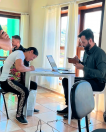 Imagem de uma sala com várias janelas amplas. No lado direito da foto está um homem da equipe da Defensoria olhando o celular. Ele está sentado em frente a uma mesa de plástico com um computador em cima. Do outro lado da mesa está uma mulher assinando um papel. Ambos são pessoas brancas. 