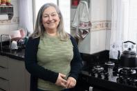Mulher branca de meia idade, cabelos grisalhos. Ela está na cozinha de sua casa, ao lado do fogão e sorri. Ela usa camiseta verde musgo e um casaco preto. 