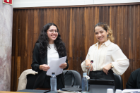Foto de duas mulheres brancas com roupas formais sorrindo. Uma tem cabelos escuros e usa óculos e segura um papel, a outra é loira. 
