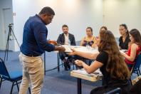 A banca está sentada na mesa, enquanto um homem negro de costas entrega um papel à moça que está mais a frente. 