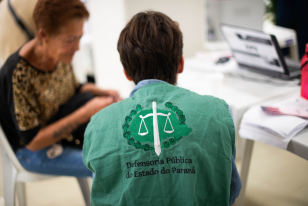 Imagem que mostra uma pessoa de costas, vestindo um colete verde com o logo da Defensoria Pública do Estado do Paraná. Ela está em atendimento a uma outra pessoa, que aparece em segundo plano, de forma desfocada. Diante delas está uma mesa com um notebook e alguns papéis. 