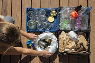 Imagem vista de cima para baixo e que mostra uma pessoa sobre um piso de madeira, organizando materiais para reciclagem. À sua frente, cestos plásticos separam os itens: um azul com potes de vidro, um preto com garrafas plásticas, outro com papel e papelão, e uma sacola com latas de metal.