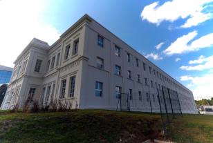 Imagem que mostra o edifício do Fórum Criminal do Ahú, em Curitiba, de cor clara e arquitetura clássica, visto de um ângulo baixo, o que o torna imponente. O prédio está situado sobre um pequeno barranco de grama, com uma grade de metal à sua frente. Ao fundo, o céu está azul com nuvens brancas e esparsas.