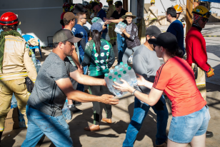 Imagem que mostra o recebimento de doações em Rio Bonito do Iguaçu. Em primeiro plano, um homem de camiseta cinza entrega um fardo grande de garrafas de água para uma mulher de camiseta vermelha. Ao fundo, voluntários e pessoas com uniformes de resgate participam da distribuição.