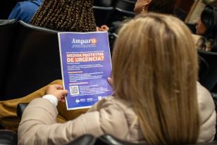 Mulher vista de costas em um auditório segura um panfleto roxo informativo do projeto Ampara da DPE-PR. O material destaca a pergunta "Medida Protetiva de Urgência?" em letras laranjas e traz um QR code com orientações para mulheres em situação de violência doméstica.