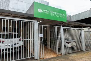 Imagem que mostra a fachada da sede da Defensoria Pública em Cascavel. O local apresenta uma grande placa verde com o logotipo da DPE-PR na parte superior e um portão de grades metálicas brancas na entrada, abrigando dois carros brancos estacionados na garagem.