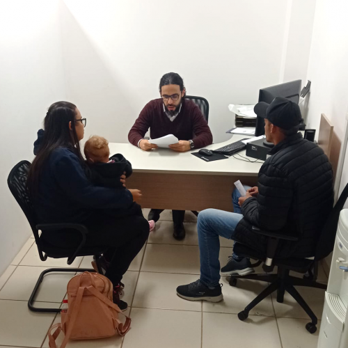 Imagem do defensor Fernando atendendo um homem e uma mulher com bebê no colo, em uma sala.