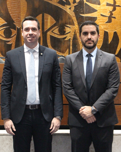 Foto do defensor público-geral, Matheus Munhoz, e do defensor público, Rafael de Matos. Eles estão em frente a um quadro marrom, em pé e de terno. O Matheus está sorrindo.