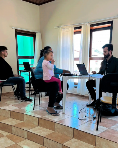 Imagem do mesmo homem atendendo, do outro lado da mesa está uma mulher com uma criança no colo. Não aparece o rosto delas.