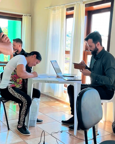 Imagem de uma sala com várias janelas amplas. No lado direito da foto está um homem da equipe da Defensoria olhando o celular. Ele está sentado em frente a uma mesa de plástico com um computador em cima. Do outro lado da mesa está uma mulher assinando um papel. Ambos são pessoas brancas.