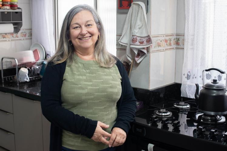 Mulher branca de meia idade, cabelos grisalhos. Ela está na cozinha de sua casa, ao lado do fogão e sorri. Ela usa camiseta verde musgo e um casaco preto.