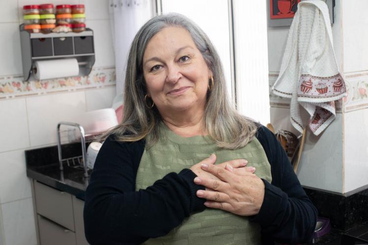Mulher branca de meia idade, cabelos grisalhos. Ela está na cozinha e sorri timidamente com as mãos no coração.