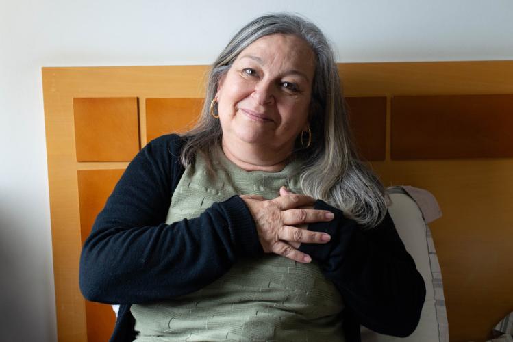 Mulher branca de meia idade, cabelos grisalhos, está sentada com as costas encostadas na cabeceira da cama. Ela olha para a câmera e sorri com as duas mãos sobre o coração. Ela usa camiseta verde musgo e um casaco preto.