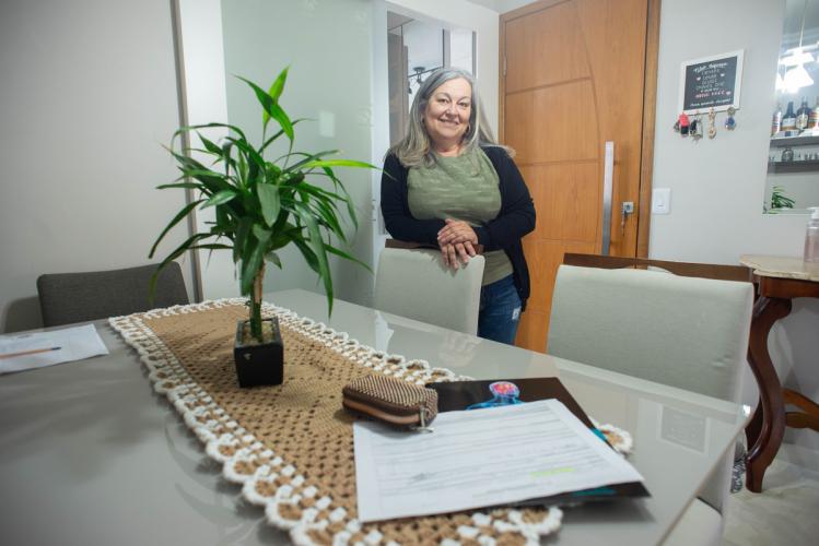Mulher branca de meia idade, cabelos grisalhos. Ela está atrás da cadeira da sala de jantar, em pé, apoiada sobre a cadeira. Na mesa, está um papel e um vaso. Ela sorri para a câmera.