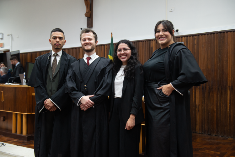 Quatro pessoas, dois homens e duas mulheres, posam no tribunal. Os dois homens estão à esquerda, de terno, gravata e toga preta; um tem cabelo curto e barba clara. À direita, uma mulher com toga e vestido preto sorri, e outra com blazer e camisa branca. Ao fundo, uma parede de madeira e uma bandeira do Brasil.